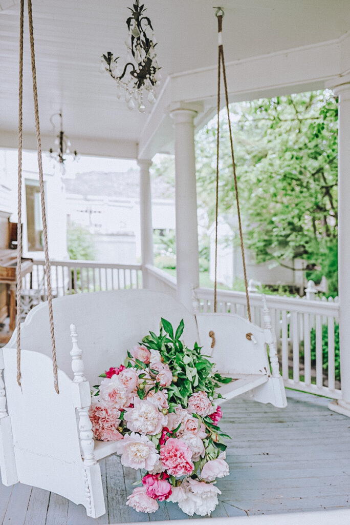 A beautiful DIY porch swing bench made out of an old headboard and footboard of an old bed. It has a chandelier above it and peonies sitting on the seat. 