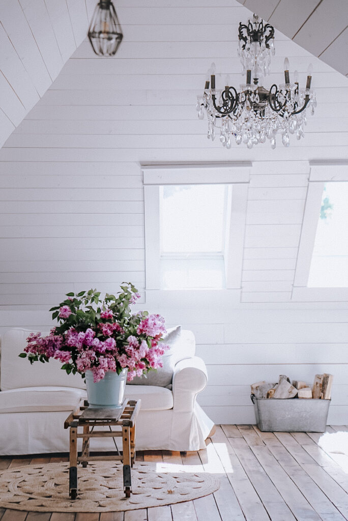 A large vintage chandelier hanging in an attic space with a sofa and a bucket full of beautiful purple lilacs .