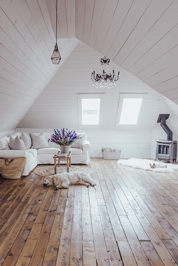 Beautiful attic space that's been finished into a family room with white shiplap walls and floors, a freestanding black fireplace, and a chandelier. 