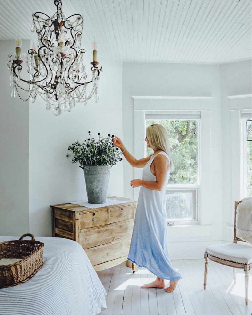 Deborah McDonald from B Vintage Style in her bedroom with a large Italian vintage French chandelier, a raw wood dresser, linen bedding, and an antique French chair. 