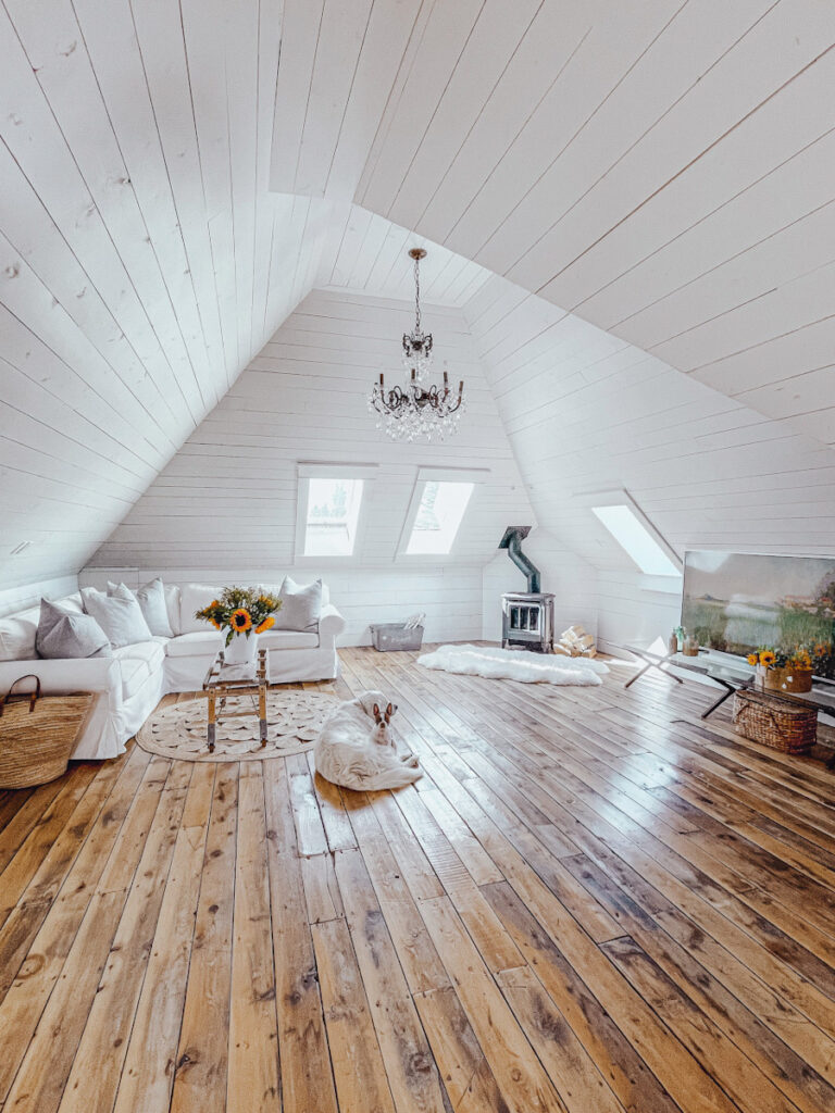 A beautiful attic space that's been converted into a family room with a fireplace, a large white sofa, a frame television, and a French chandelier. 