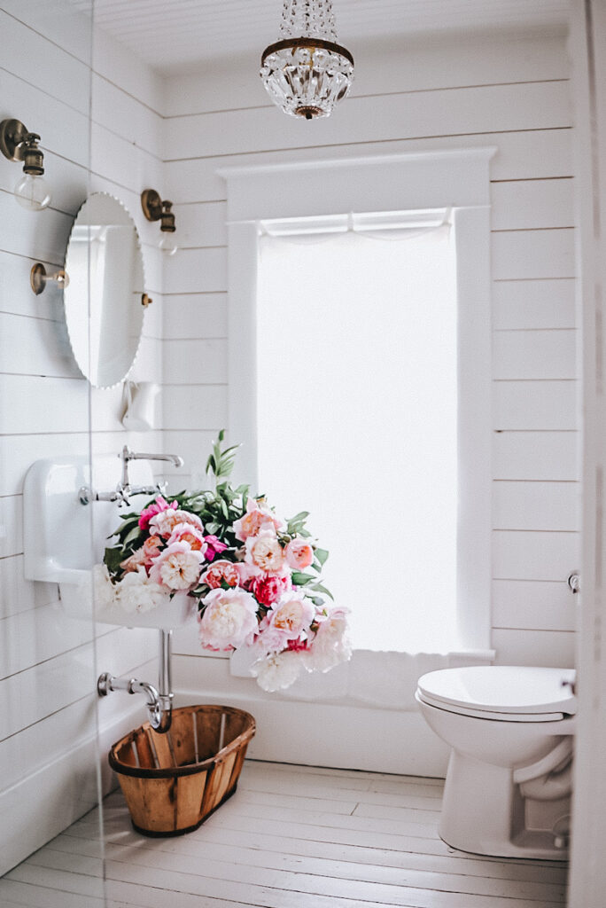 A beautiful white farmhouse bathroom with shiplap walls and a massive bundle of peonies in the sink and a French antique chandelier.