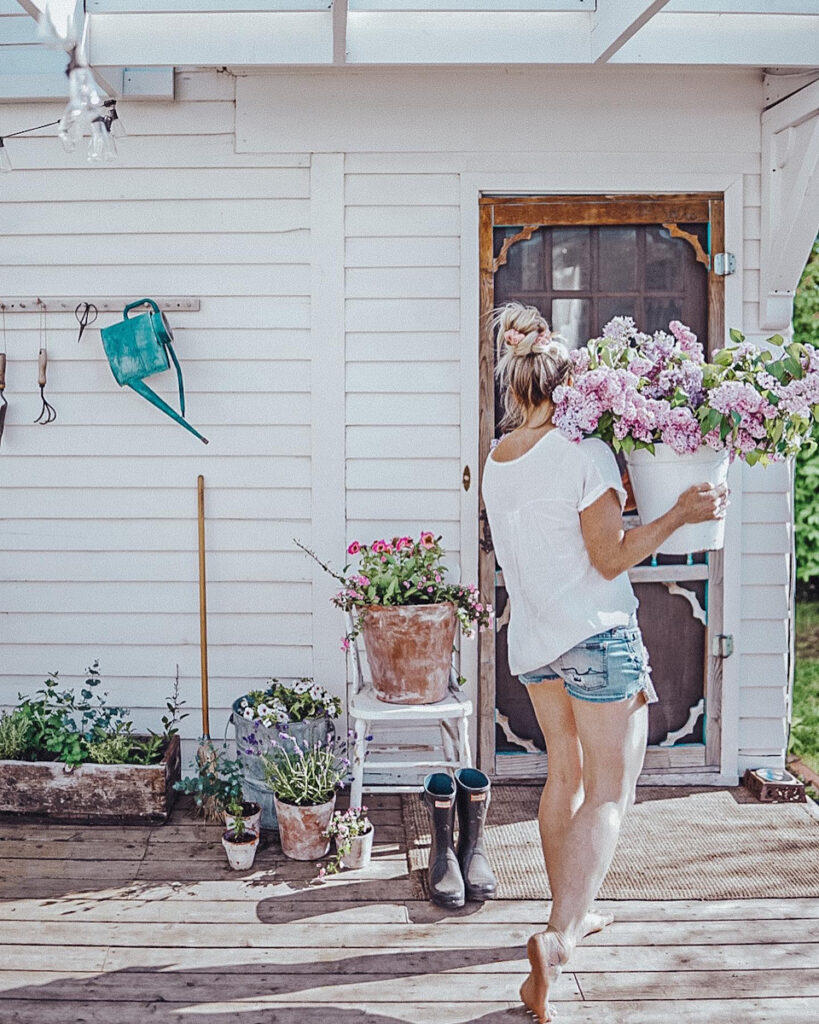 Deborah from B Vintage Style carrying a bucket of lilacs on her back patio with terracotta pots and planters.