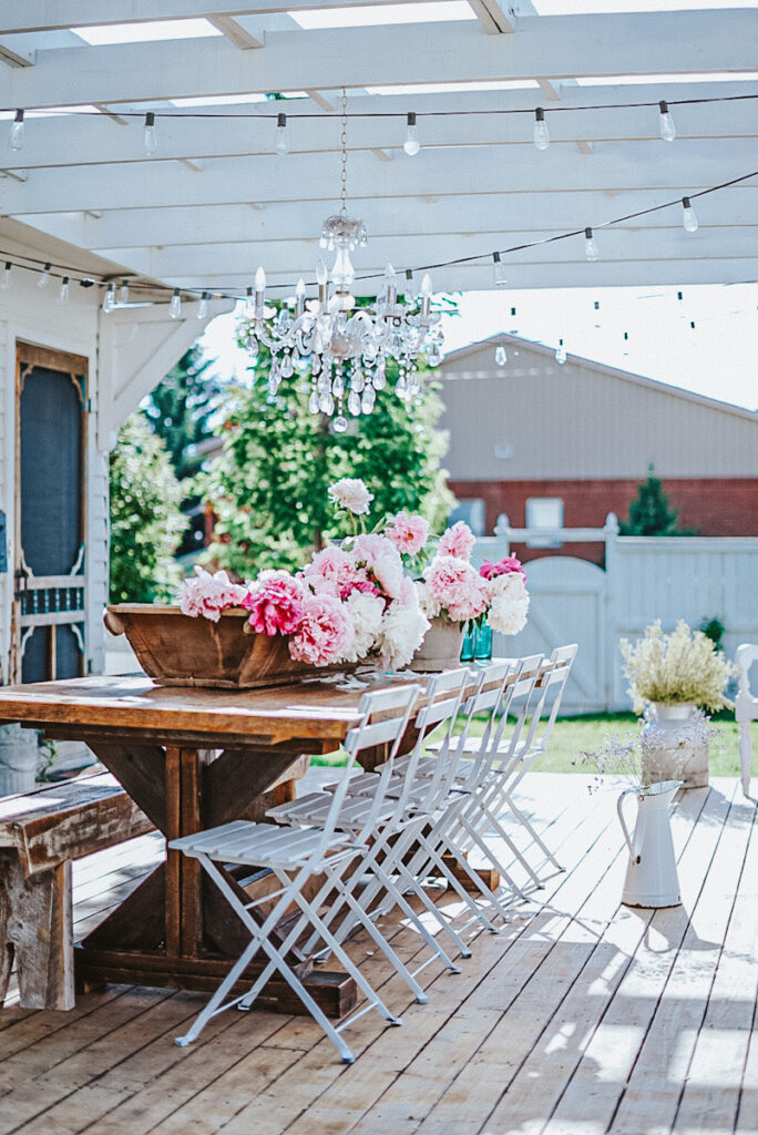 A farmhouse table on an outdoor patio with French cafe chairs and a crystal chandelier hanging above.