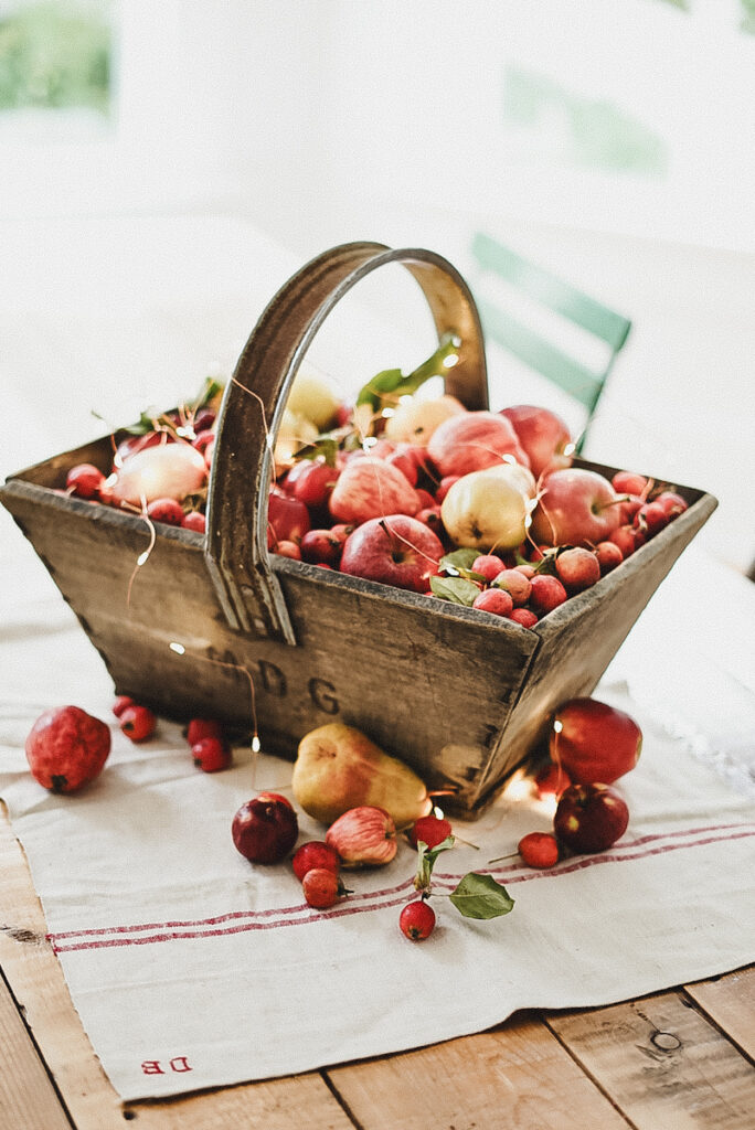 Wood basket of apples sits on a table with twinkle lights over top of them, along with a few apples and twinkle lights out of the basket sitting beside it.