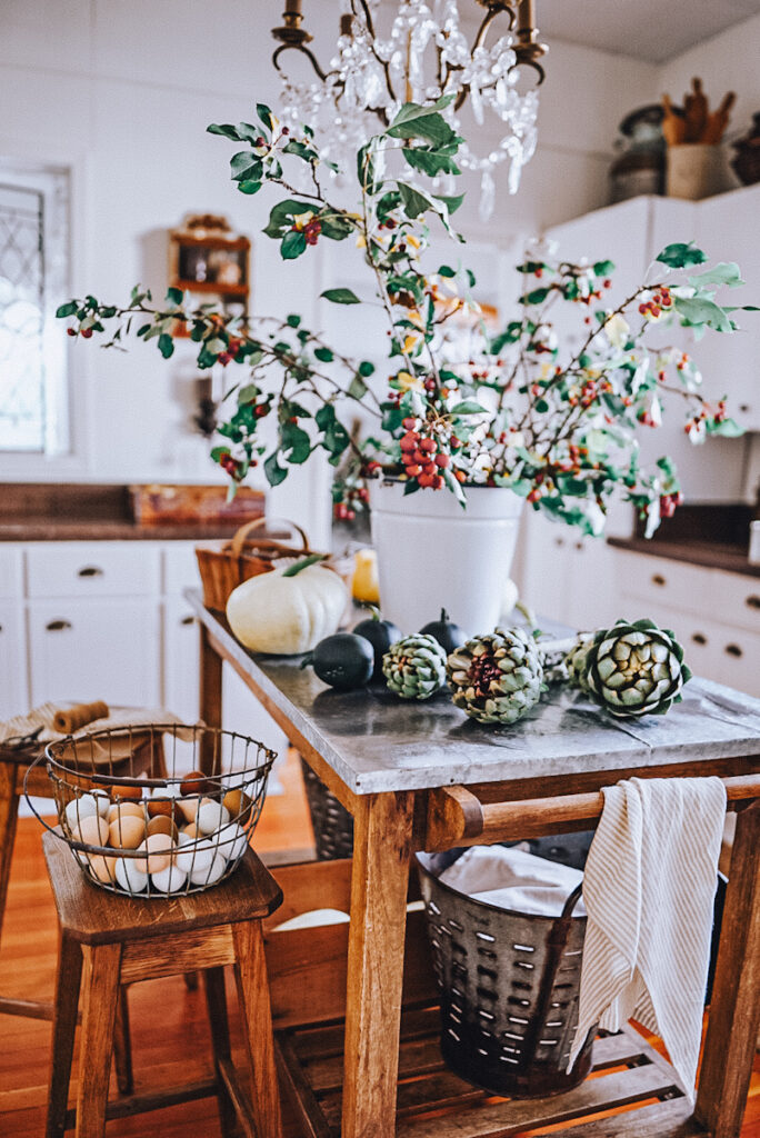 Artichokes sit on the table along with pumpkins in front of a white bucket with crab apples, there is a basket of eggs beside the table on a stool