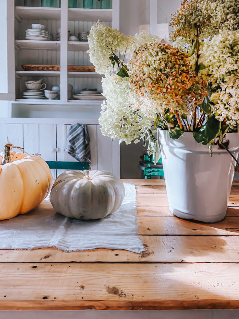 Two pumpkins sit on a table along with a bucket of flowers. Behind the table is a cabinet.