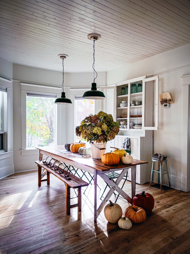 Pumpkins sit on a table along with a white bucket filled with flowers.