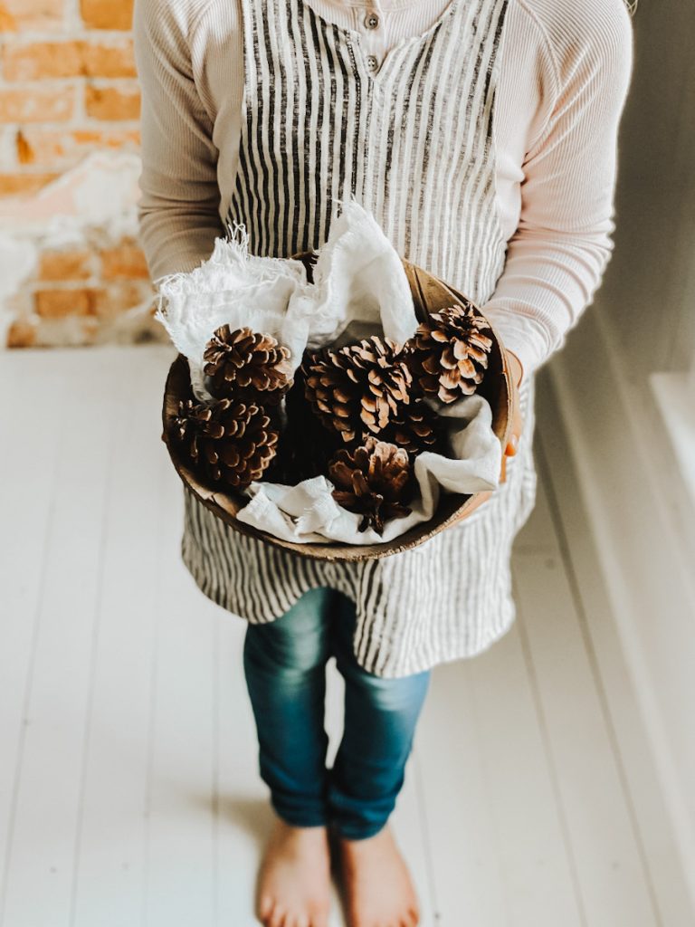 young girl in a linen apron holding an antique wooden bowl filled with pine cones for winter decorations