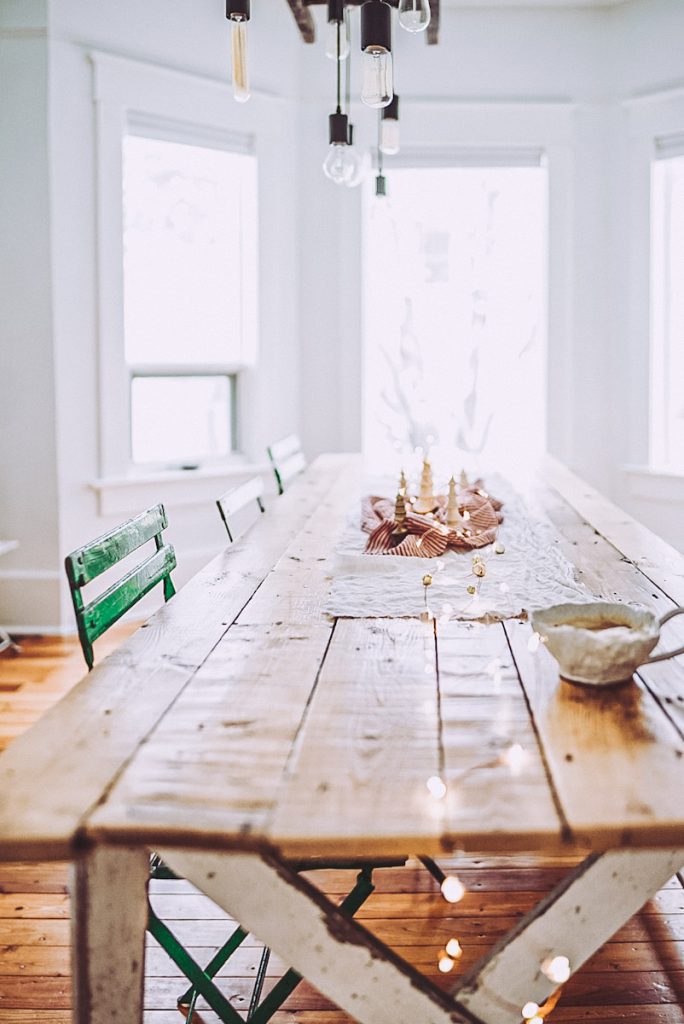 A farmhouse dining table that is decorated with twinkle lights, linen and tiny wooden trees for winter