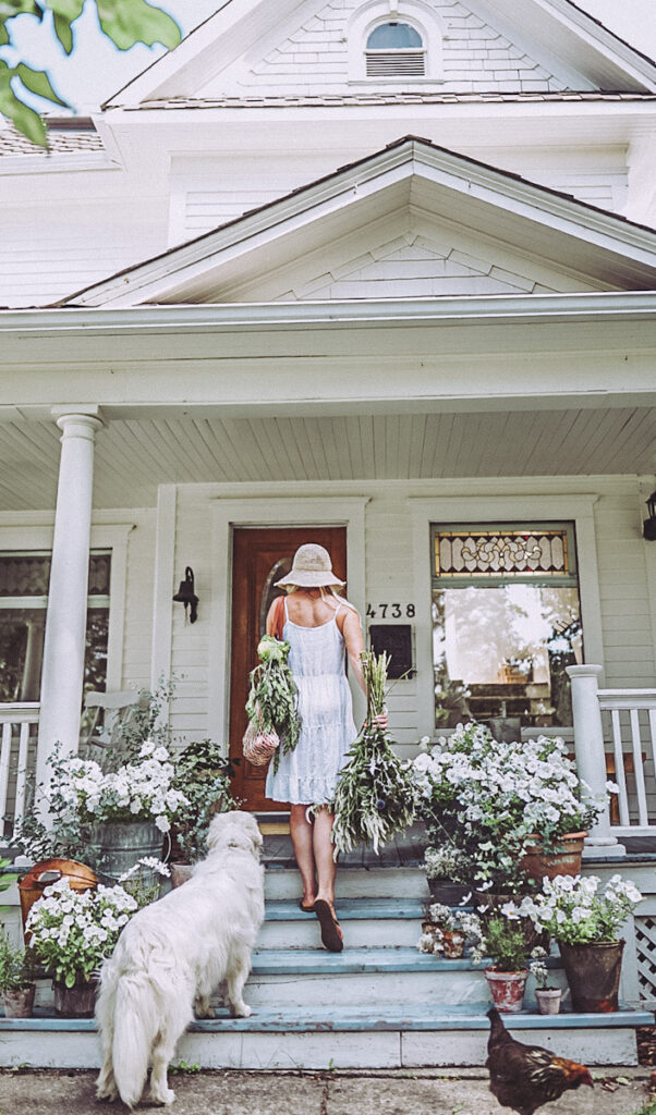 Deborah from Be Vintage Style, walking up her front porch steps that are full of flowers and planters and DIY Aged Terra Cotta Pots 