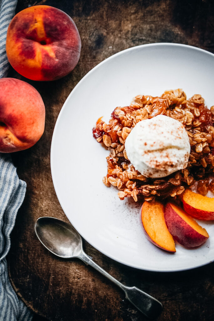 Yummy peach crumble that has vanilla ice cream on top and sliced fresh peaches beside it, with a silver spoon on a white plate.