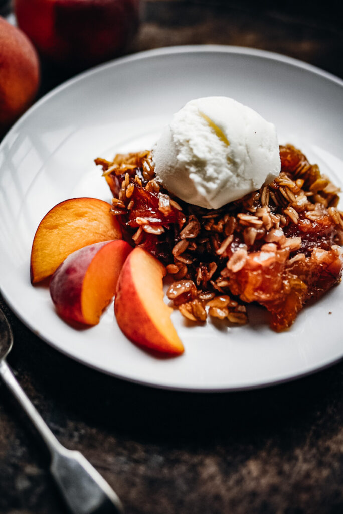 Peach crumble with a scoop of vanilla ice cream on a white plate and slices of fresh peaches beside it.