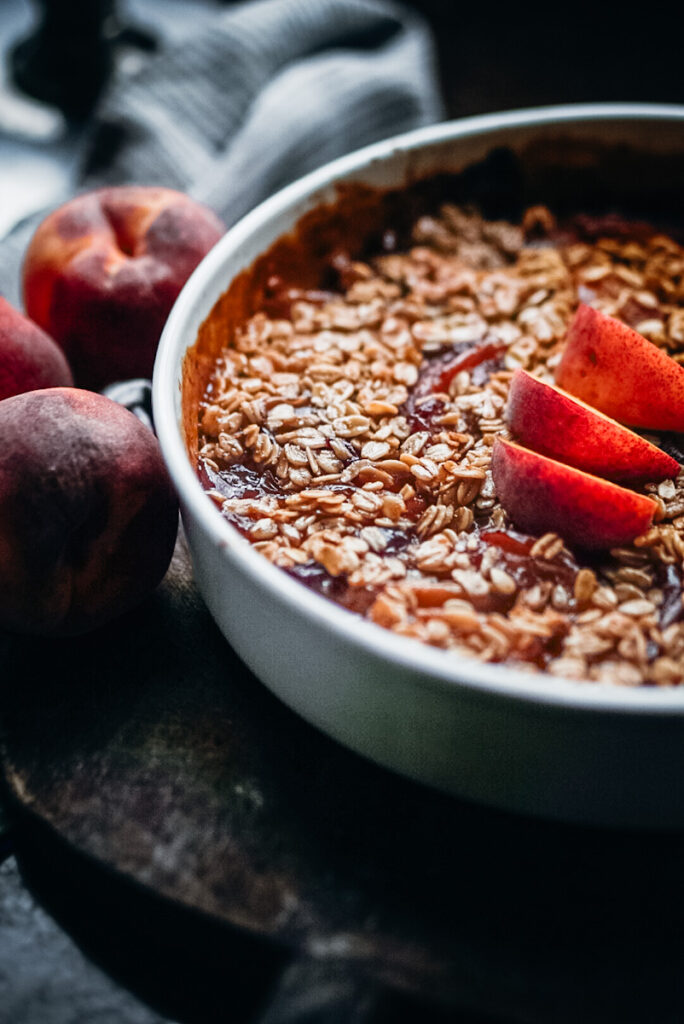 Peach crumble that's just been finished in a white vintage dish with fresh sliced peaches on top.