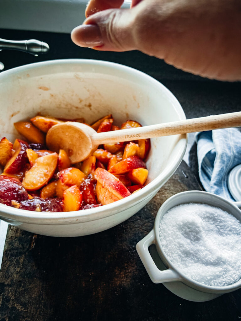 Fresh peaches that are tossed with sugar and cinnamon, ready to be made into a crumble in a big bowl with a wooden spoon.