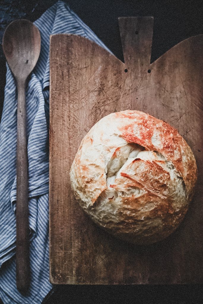 A large loaf of Artisan bread on an antique cutting board with a linen towel and wooden spoon beside it.