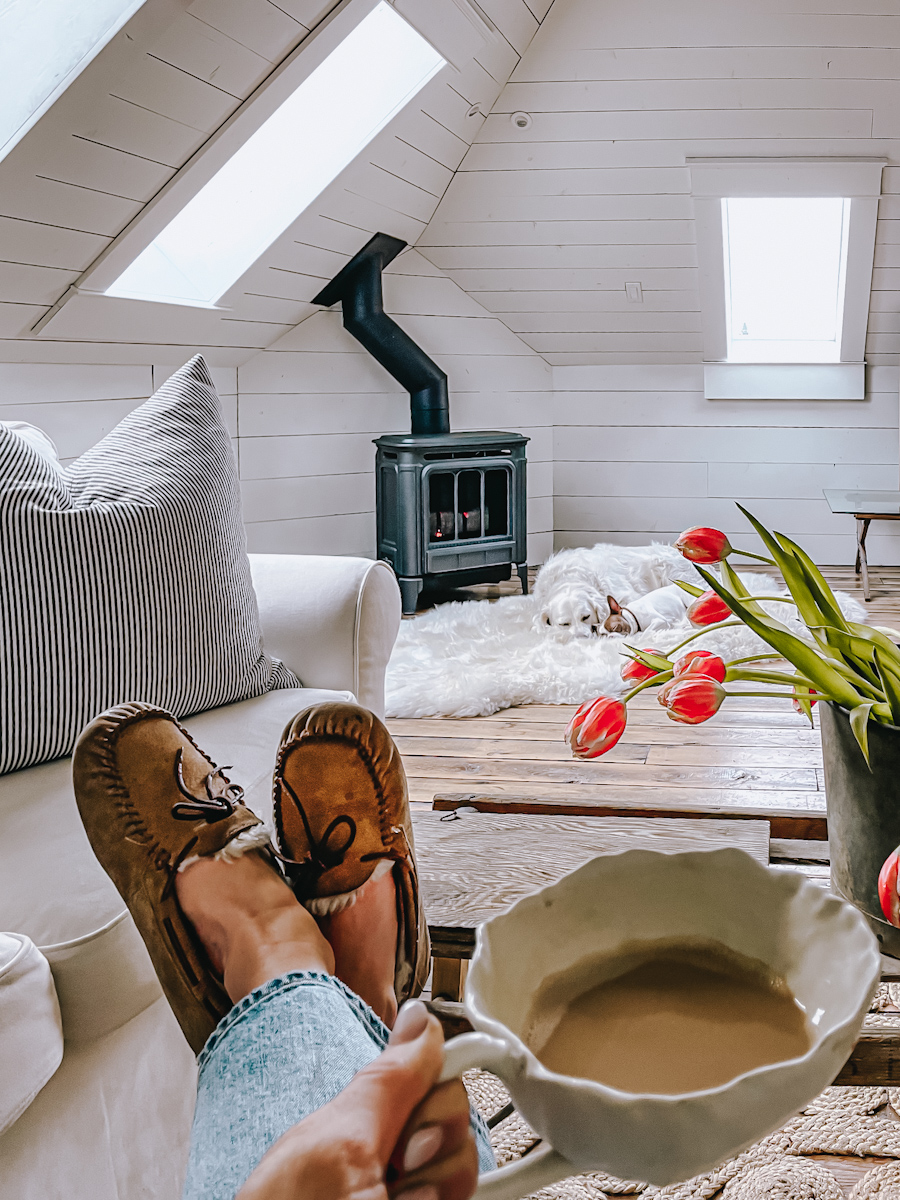 A cozy Hygge style fireplace with a sheep skin rug in front and two dogs sleeping. A girl with her feet up on the table, wearing slippers and drinking coffee.
