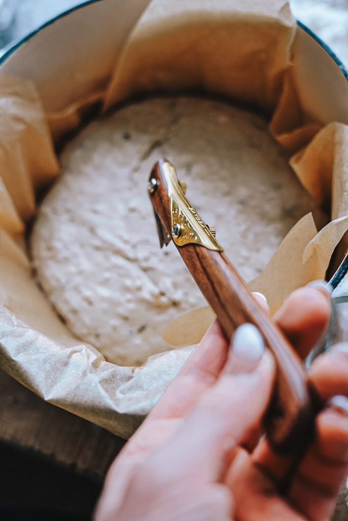Using a bread lame to make a design on a loaf of bread in a Dutch oven with parchment paper.