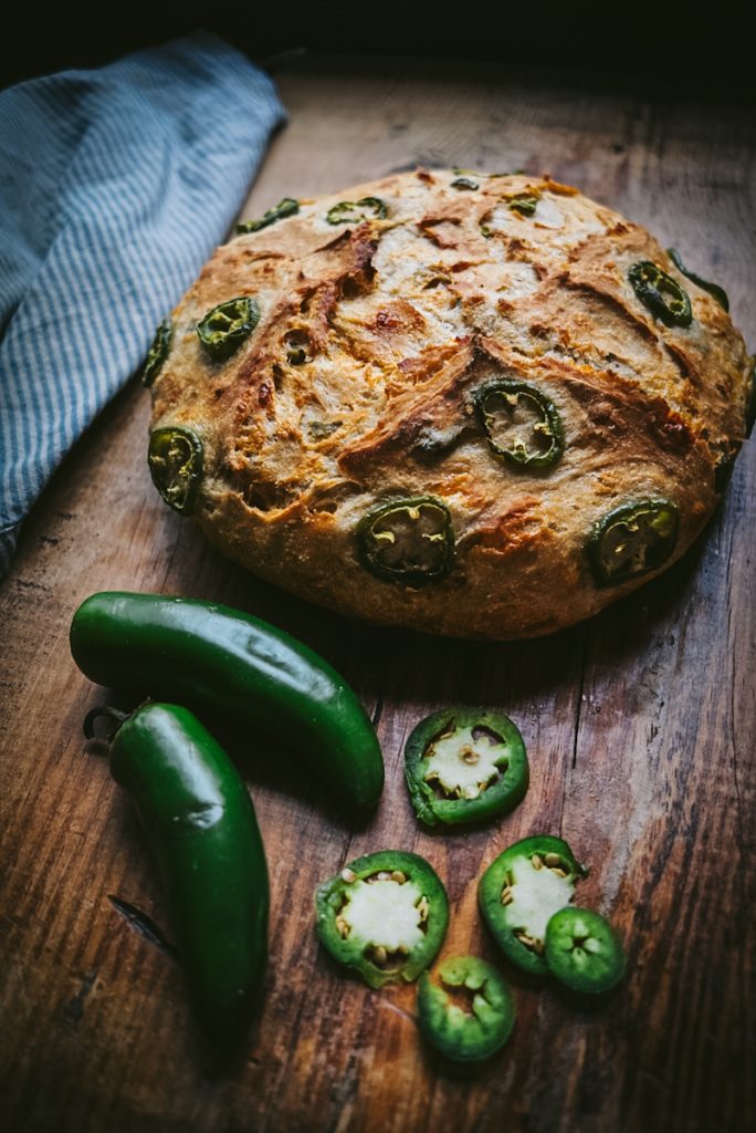 A fresh loaf of jalapeno cheese bread on a vintage cutting board beside some sliced jalapenos and jalapeno peppers.