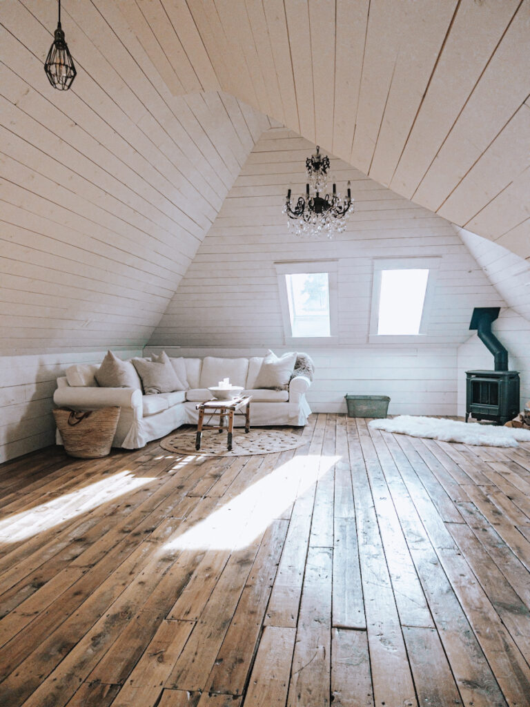 Beautiful attic space that is used as a familyroom with white shiplap walls, a  chandelier, a fireplace, and a sectional.