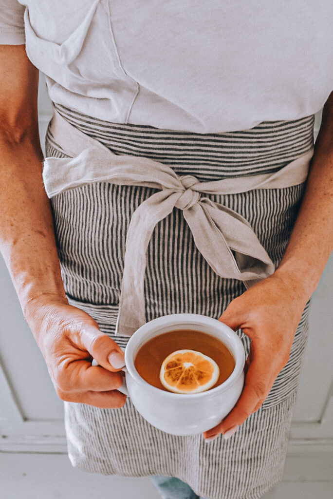 A girl holding a cup of homemade apple cider with an orange slice garnishing the top, wearing a linen apron.