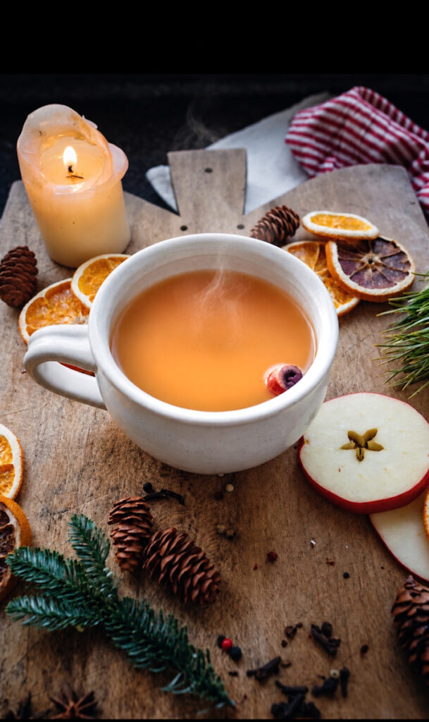 A delicious, steaming hot mug of hot apple cider with apple slices, orange slices, pine cones, and a candle beside it.