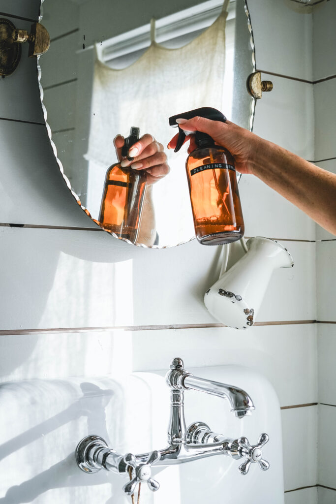 Spraying a mirror with a DIY window cleaning spray in an amber glass bottle.