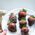 Dark chocolate-dipped strawberries on a white plate that look delicious with sprinkles, and nuts.