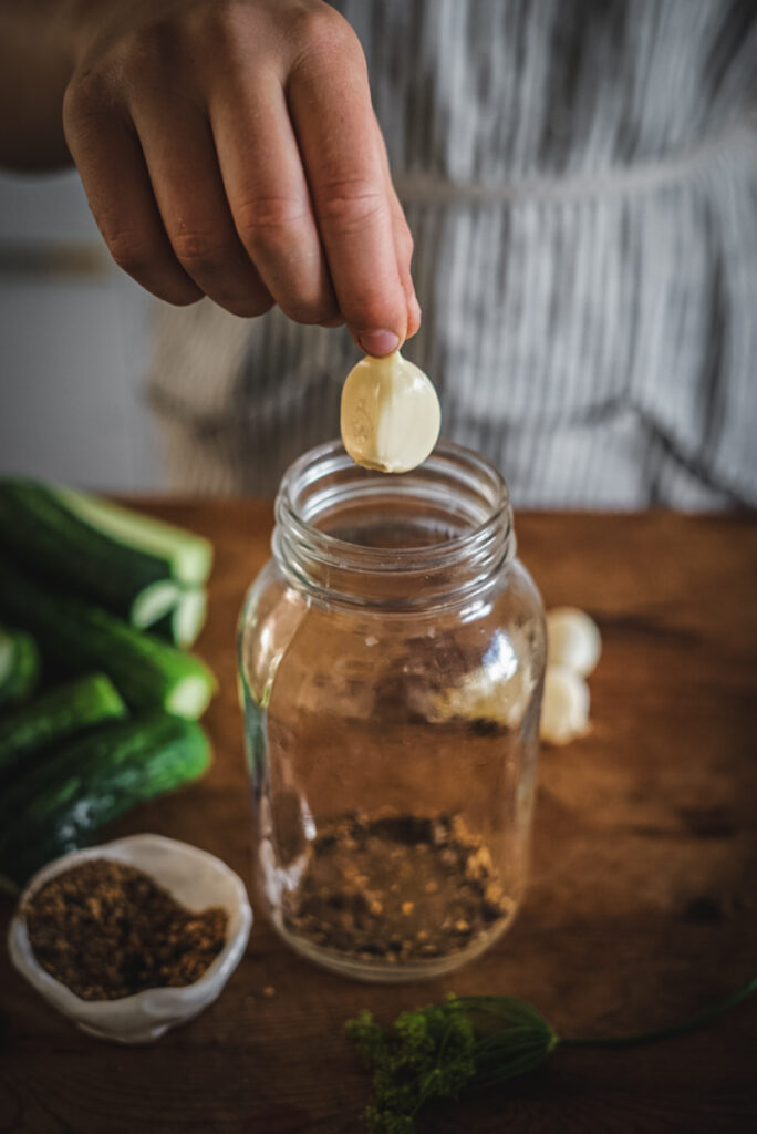 Adding garlic and pickling spices to jars to make dill pickles.