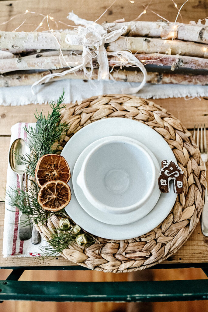 A beautiful white dining setting up for Christmas with dried orange slices, fresh juniper, sprigs, and a gingerbread house.