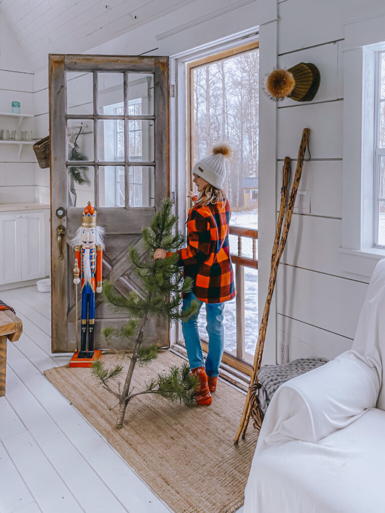 A young girl carrying a Christmas tree out a cabin door, with a tall nutcracker holding it open.