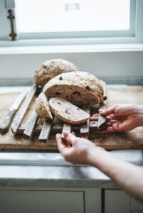 Fresh loaf of cranberry bread being placed in front of a large window on an antique cutting board.