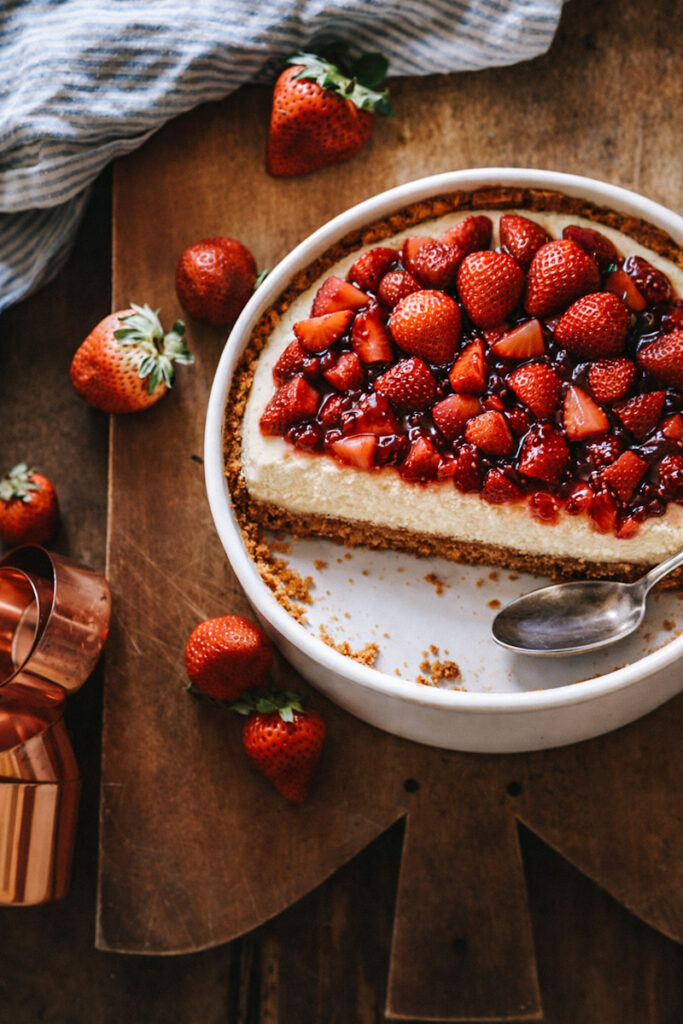 A strawberry cheesecake that has been partially eaten in a white ceramic dish on a wooden cutting board surrounded by extra strawberries. 