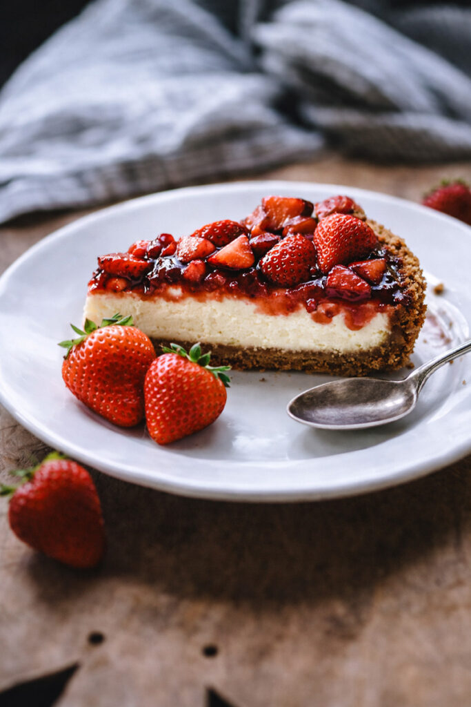 A slice of Philadelphia strawberry cheesecake on a white plate with a spoon and a few strawberries on the side. 