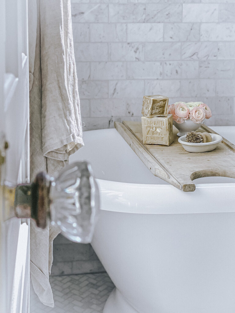 A beautiful claw foot tub with a shower caddy that has Marseille soap, fresh ranunculus, and a scrub brush. 