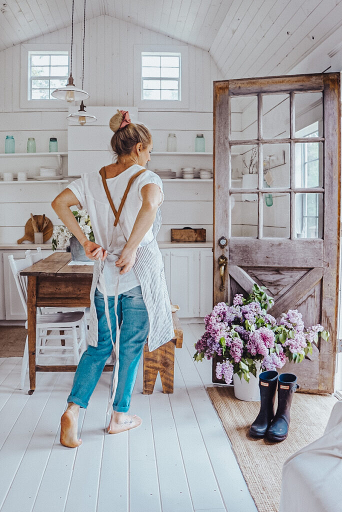 A tiny house shed cabin with a girl walking through it and a big bucket of lilacs. 