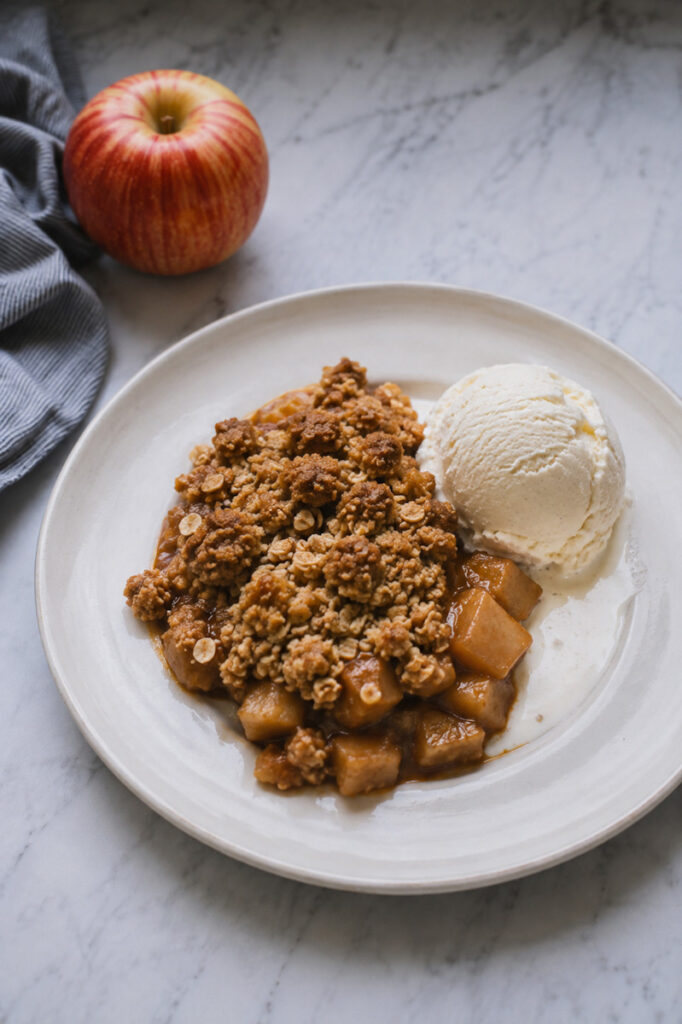 Serving a warm buttery apple crumble with vanilla ice cream and an apple beside the dish.