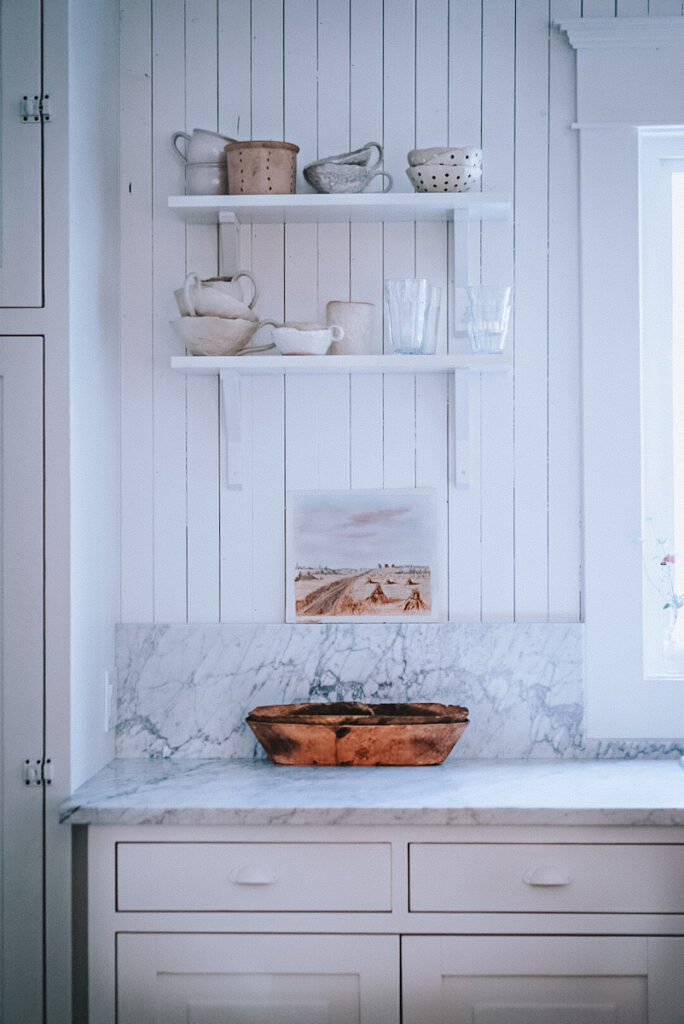 A beautiful vintage piece of watercolor autumn artwork that's been thrifted and showcased in a kitchen below shelves of pottery.