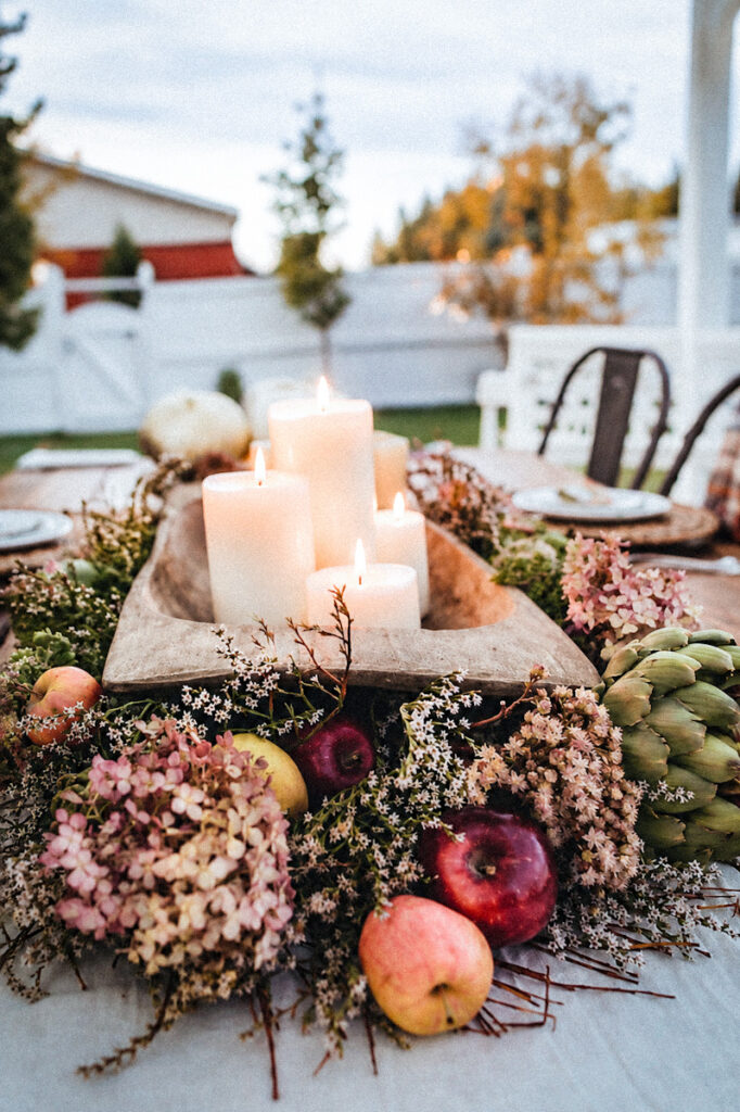 A stunning centerpiece on an outdoor dining table with fall foliage, like apples, artichokes, dried hydrangeas, and willow, with lit candles.