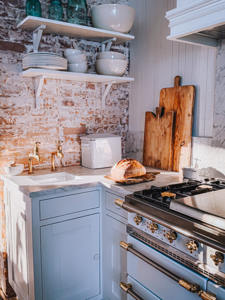 An exposed brick wall in a kitchen that has open shelves, antique cutting boards, and a loaf of bread in front of a beautiful French Lacanche stove.
