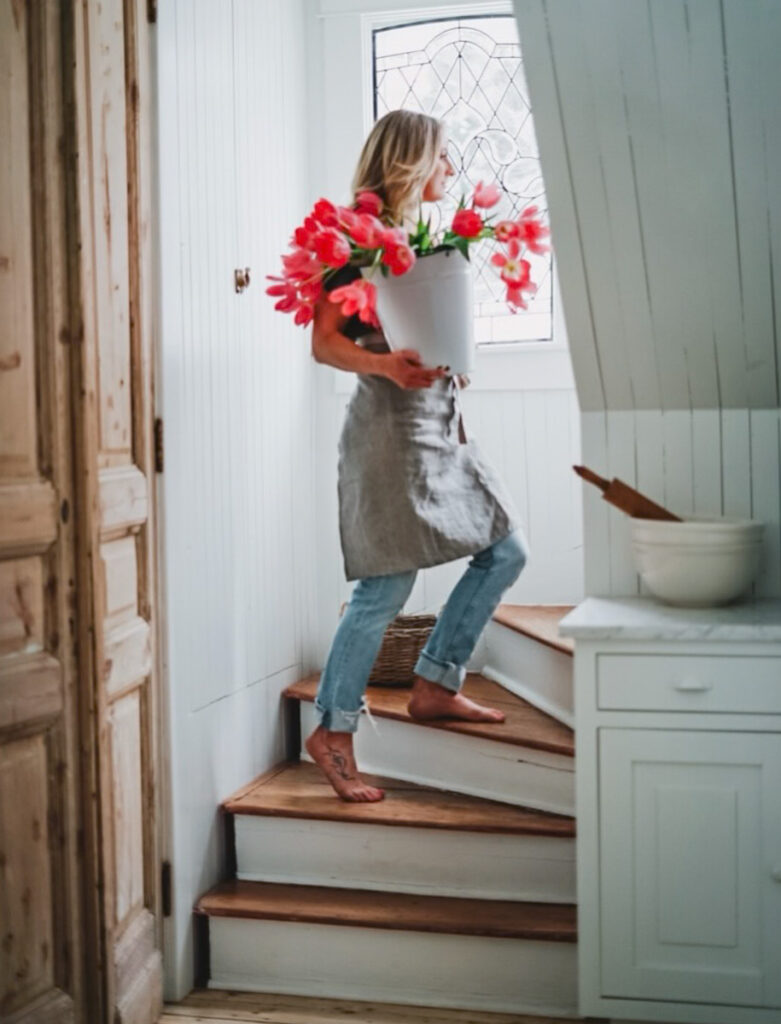 Deborah from B Vintage Style walking up a staircase carrying a bucket of tulips and wearing a linen apron.