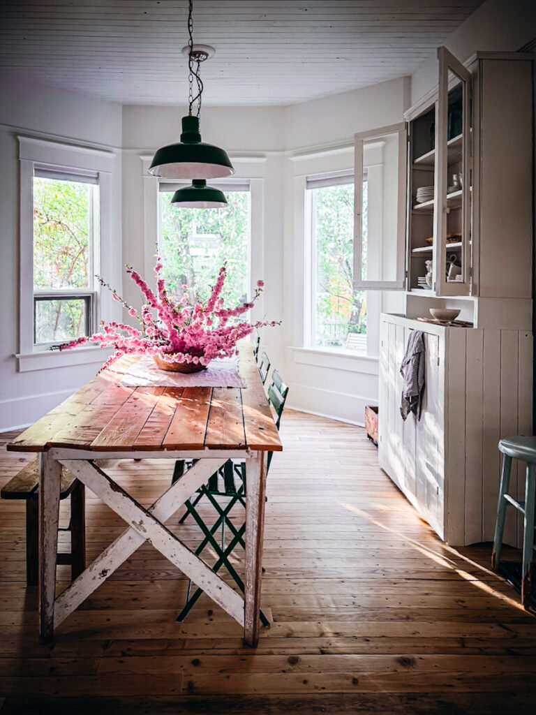 A beautiful living room space with a farmhouse antique table, beautiful lighting, a white kitchen hutch, and flowers on the table.