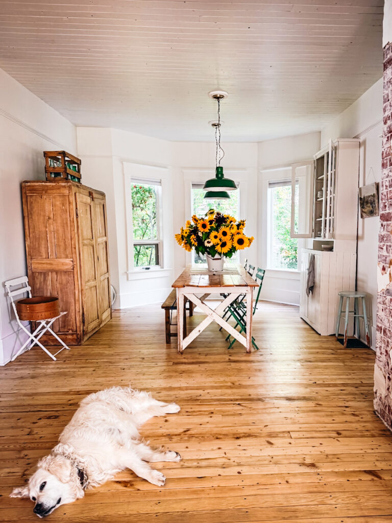 A calm living room space with a bay window, antique rustic furniture, a farmhouse table with a massive bouquet of sunflowers on it, and a dog lying in front.
