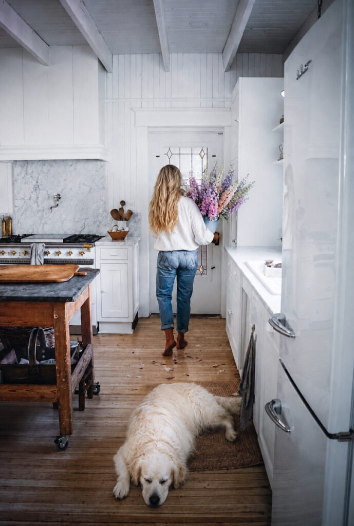 Deborah from B Vintage Style walking through her kitchen, carrying a bucket of larkspur, wearing a white shirt and jeans, with her dog lying on the floor in front of her.