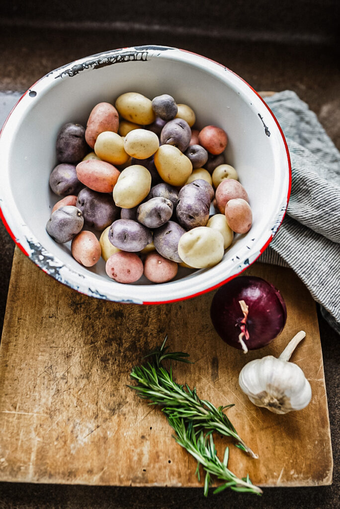 A bowl full of colorful baby potatoes on a cutting board with onion, garlic, and rosemary to make roast beef.